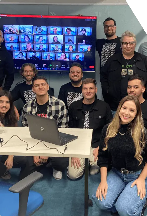 Turma do Vem Ser DBC reunida presencialmente com uma tv no fundo com os participantes de forma remota